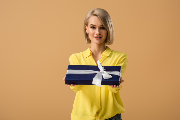 Pretty blonde young woman showing gift box with ribbon isolated on beige - Фото, изображение