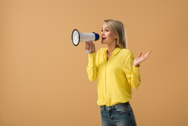 Smiling blonde woman in yellow shirt screaming in megaphone isolated on beige - Фото, изображение