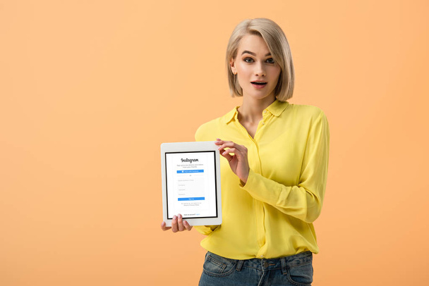 Surprised girl in yellow shirt holding digital tablet with instagram app on screen isolated on orange - Φωτογραφία, εικόνα