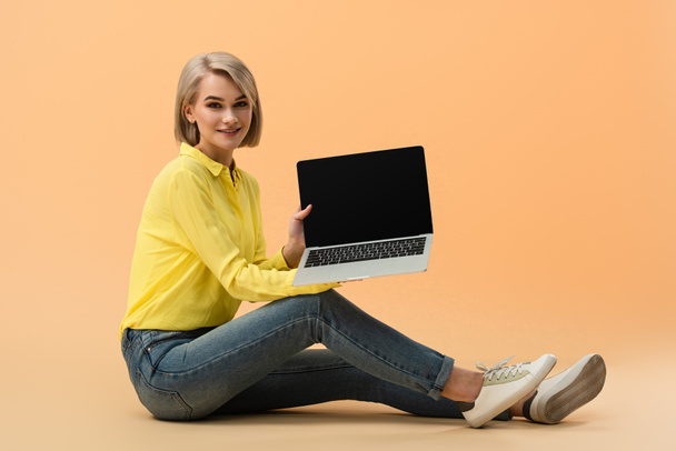 Uśmiechnięta Blondynka w dżinsy Wyświetlono laptop rezygnować pusty ekran, siedząc na pomarańczowym tle - Zdjęcie, obraz
