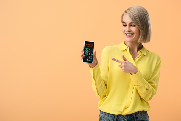 Smiling woman pointing with finger at smartphone with marketing analysis app on screen isolated on orange - Photo, Image