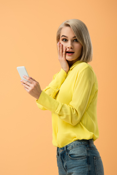 Stylish surprised blonde girl holding smartphone isolated on orange - Foto, Bild