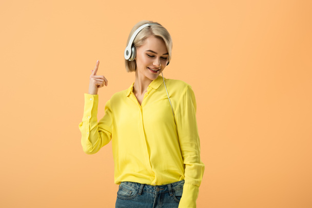 Joyful blonde woman in yellow shirt listening music in headphones isolated on orange - Foto, immagini
