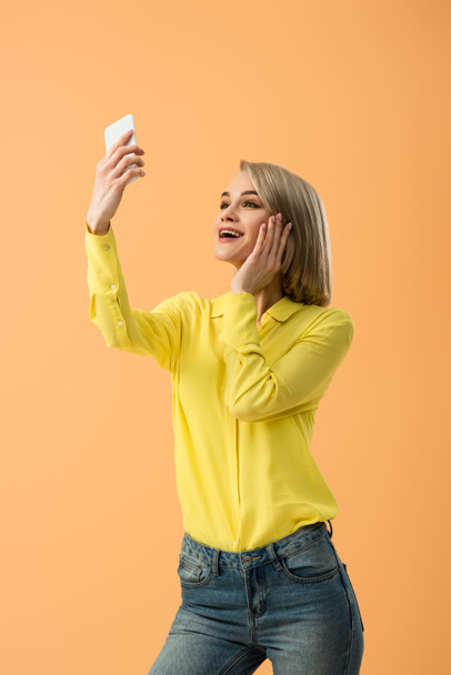 Surprised blonde woman in yellow shirt taking selfie isolated on orange - Foto, afbeelding