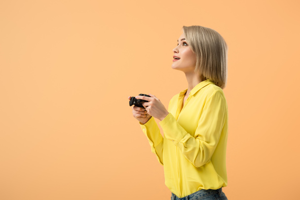 Mujer joven soñadora con camisa amarilla sosteniendo el joystick y mirando hacia arriba aislado en naranja
 - Foto, Imagen