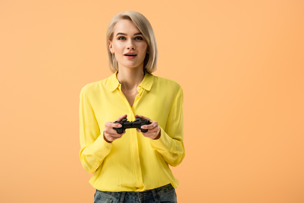 Interested blonde girl in yellow shirt holding gamepad isolated on orange - Fotoğraf, Görsel