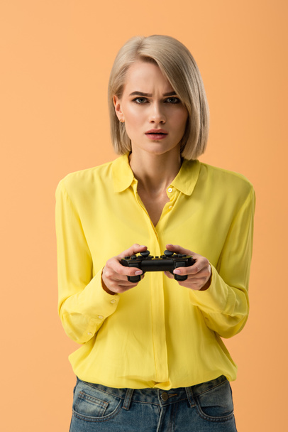 Mujer rubia preocupada en camisa amarilla sosteniendo gamepad aislado en naranja
 - Foto, Imagen