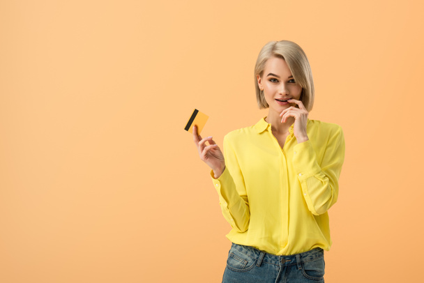 Sensual blonde woman in yellow shirt holding credit card isolated on orange - Foto, Imagen