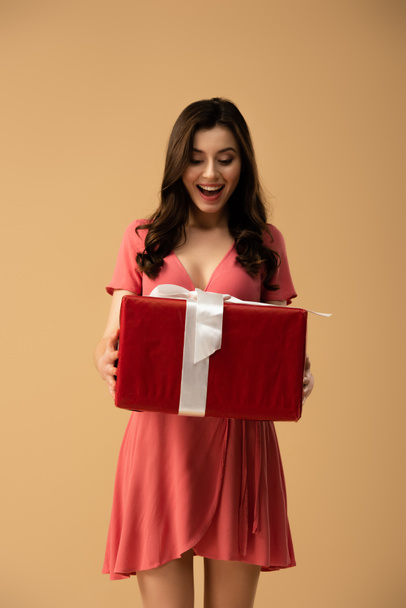 Surprised curly woman in dress holding big gift box isolated on beige - Fotoğraf, Görsel