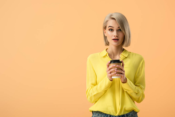 Surprised young woman in yellow shirt holding paper cup of coffee isolated on orange - Photo, image
