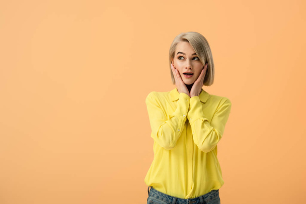 Shocked blonde woman in yellow shirt touching face and looking up isolated on orange - Фото, зображення