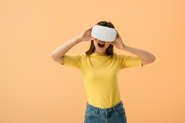 Excited brunette girl touching vr headset with open mouth isolated on orange - Φωτογραφία, εικόνα