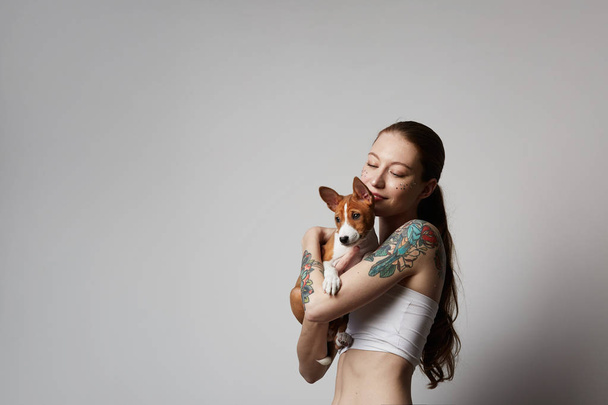 Portrait of a cute tattooed young woman hugging and kissing her little puppy basenji dog. Love between dog and owner. Isolated over white background. - Photo, Image