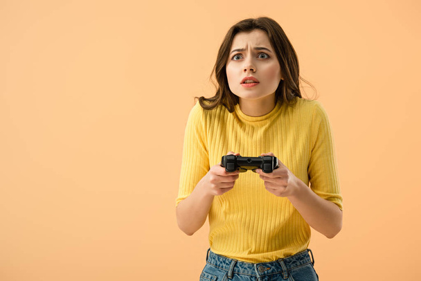 Frightened brunette young woman holding gamepad isolated on orange - Фото, изображение