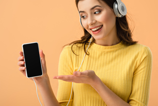 Happy brunetka ukazuje smartphone s prázdnou obrazovkou izolované na orange sluchátka - Fotografie, Obrázek