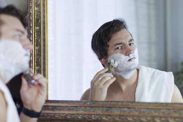 Young handsome caucasian man begins to shave with brush and foam (mirror reflection), vintage style of old barber. Thoughtful serious look, towel on the shoulder, young metrosexual cares himself. White background, indoors, copy space. - 写真・画像