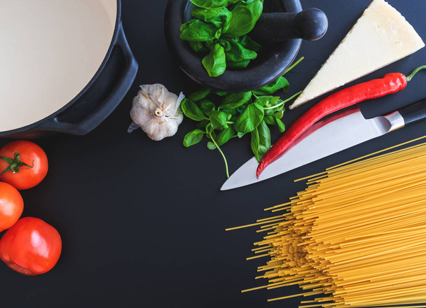 voorbereiding van Italiaanse pasta met verse basilicum in de mortel, tomaten, knoflook, peper en Parmezaanse kaas op donkere aanrecht - Foto, afbeelding