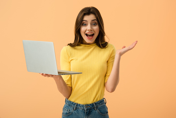 Surprised brunette girl holding laptop and looking at camera isolated on orange - Photo, Image