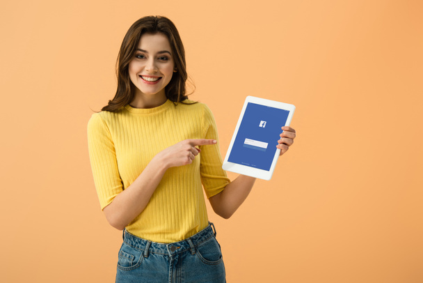 Gorgeous smiling girl pointing with finger at digital tablet with facebook app on screen isolated on orange - Fotoğraf, Görsel
