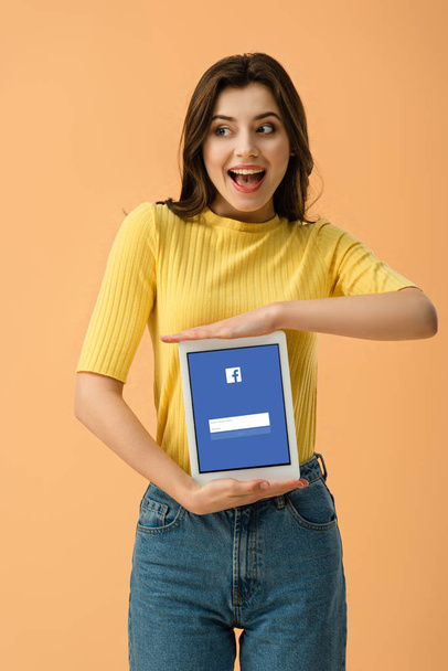 Excited brunette girl holding digital tablet with facebook app on screen isolated on orange - Foto, immagini