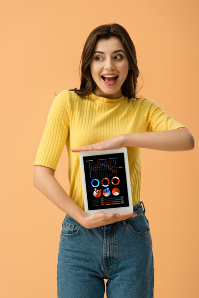 Happy brunette girl holding digital tablet with business app on screen isolated on orange - 写真・画像