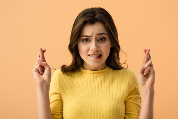 attractive brunette woman biting lips while standing with fingers crossed  isolated on orange  - Photo, Image