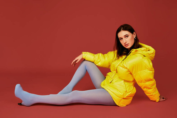 Studio portrait of young brunette woman sitting on the floor in yellow down jacket and grey blue panty hoses or stockings - Photo, Image