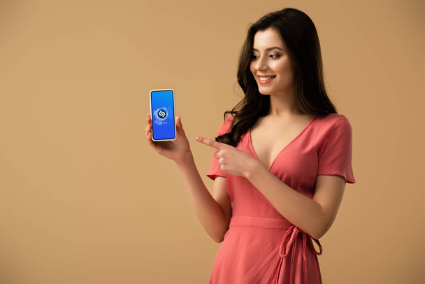 cheerful brunette girl pointing with finger at smartphone with shazam app on screen isolated on brown  - 写真・画像