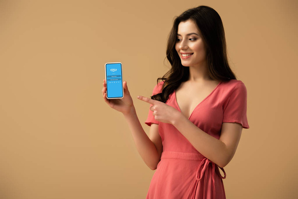 cheerful brunette girl pointing with finger at smartphone with skype app on screen isolated on brown  - Fotó, kép