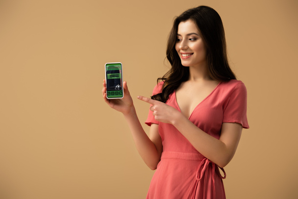 cheerful brunette girl pointing with finger at smartphone with booking app on screen isolated on brown  - Fotografie, Obrázek