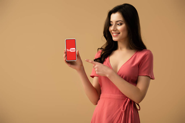 cheerful brunette girl pointing with finger at smartphone with youtube app on screen isolated on brown  - Foto, Bild