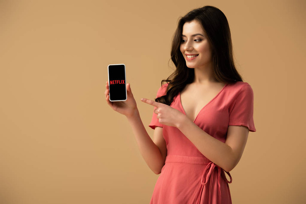 εύθυμος μελαχρινή κοπέλα, δείχνοντας με το δάχτυλο στο smartphone με εφαρμογή netflix στην οθόνη που απομονώνονται σε καφέ  - Φωτογραφία, εικόνα