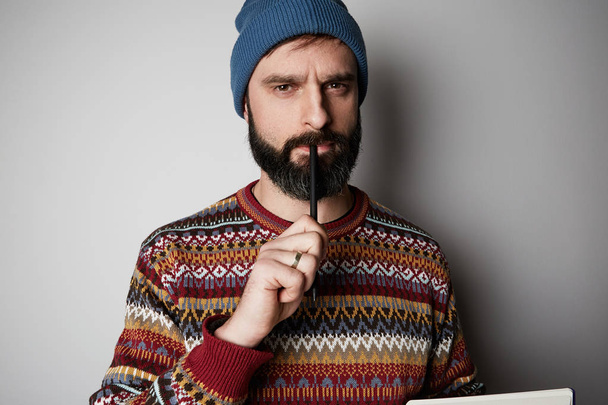 Young thoughtful bearded man in blue beanie thinking about test over empty background. - Foto, Imagem