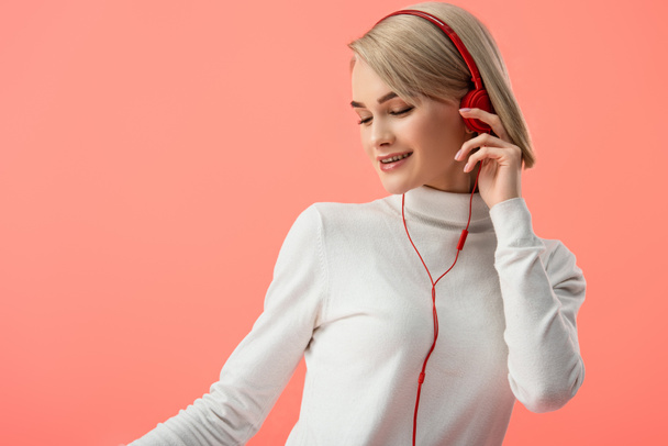 glückliche blonde junge Frau hört Musik in Kopfhörern isoliert auf rosa  - Foto, Bild