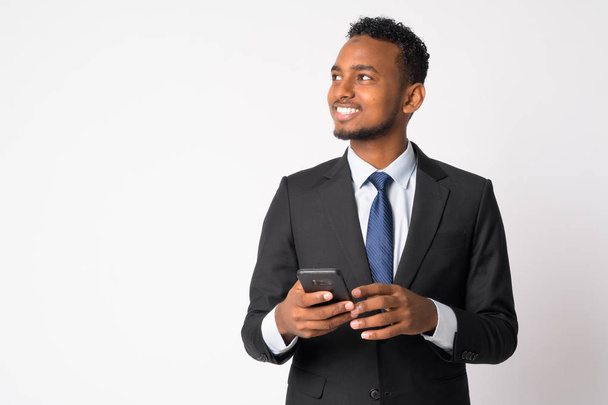 Happy young handsome African businessman thinking while using phone - Fotó, kép