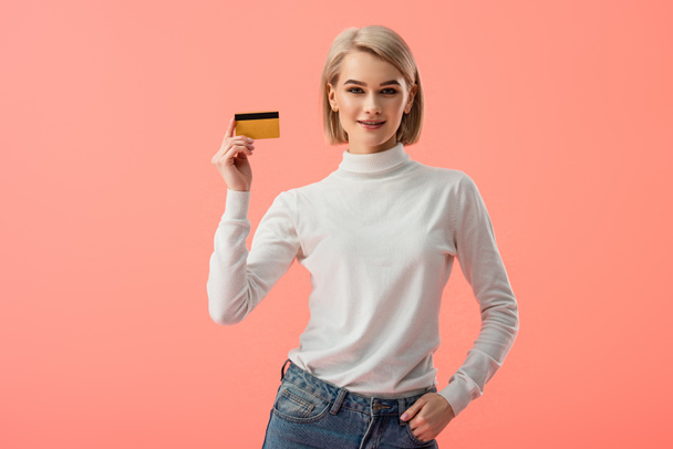 happy blonde young woman holding credit card and standing with hand in pocket isolated on pink  - Фото, зображення