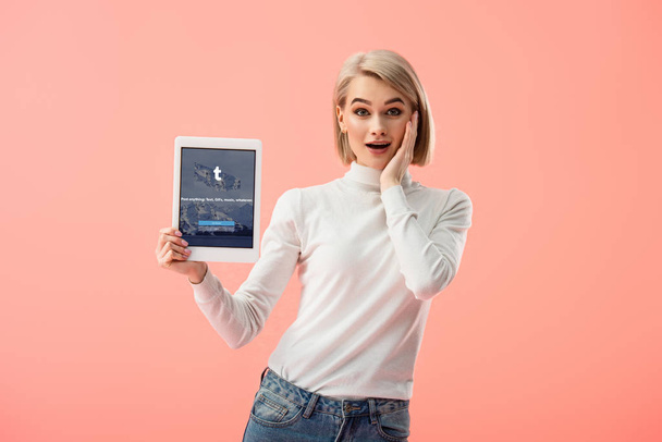 surprised blonde woman holding digital tablet with tumblr app on screen isolated on pink  - 写真・画像