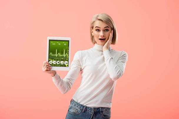 surprised blonde woman holding digital tablet with health app on screen isolated on pink  - Fotó, kép