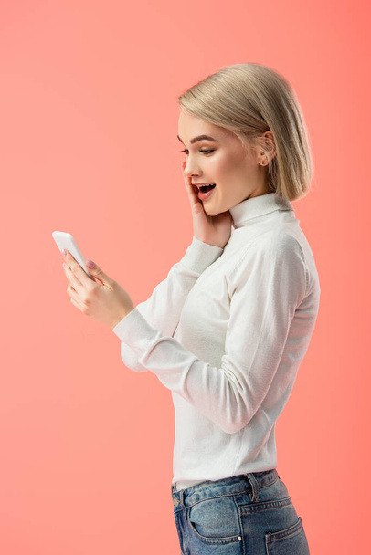 shocked blonde girl looking at smartphone isolated on pink  - Foto, Imagem