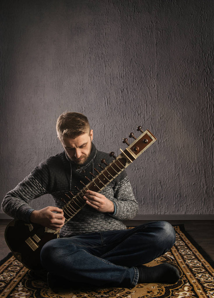 Portrait of a European man playing the sitar sitting on the carpet - Photo, image
