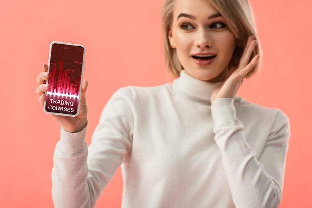 foco seletivo de mulher loira surpreso segurando smartphone com gráficos na tela isolada em rosa
  - Foto, Imagem
