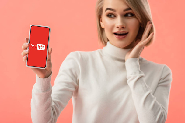 selective focus of surprised blonde woman holding smartphone with youtube app on screen isolated on pink  - Foto, immagini
