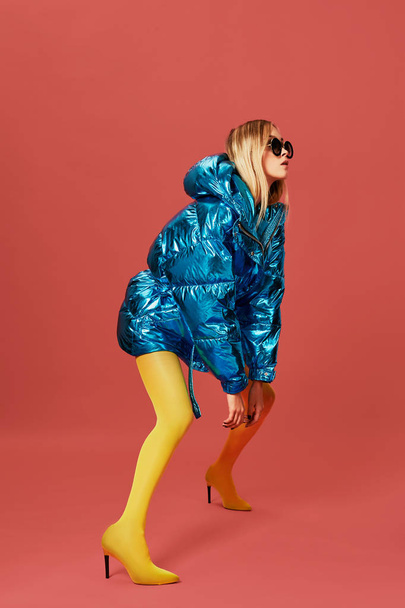 Alluring skinny young blonde woman posing in studio on red background. She is wearing blue down jacket, sunglasses, yellow stockings - Photo, Image