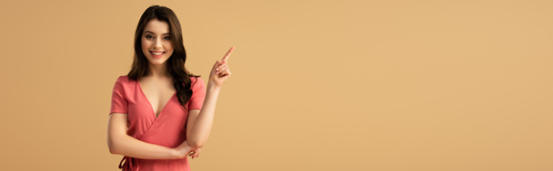 Panoramaaufnahme einer fröhlichen brünetten Frau, die mit dem Finger zeigt, während sie isoliert auf braun steht  - Foto, Bild