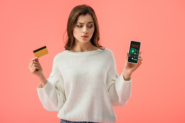 atraktivní bruneta dívka drží kreditní kartu při pohledu na smartphone s marketingové analýzy na obrazovce izolované na růžové  - Fotografie, Obrázek