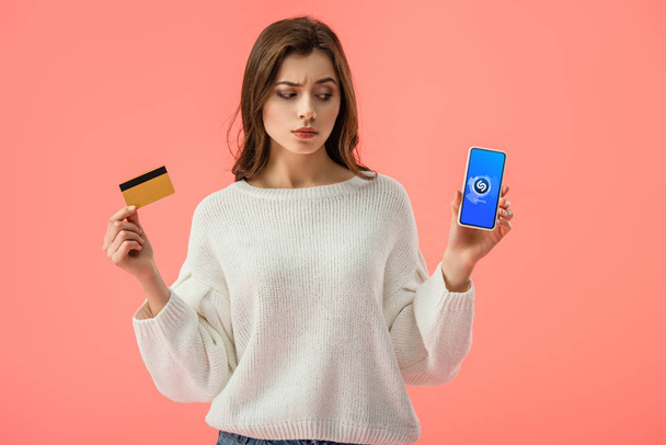 atraktivní bruneta dívka drží kreditní kartu při pohledu na smartphone s aplikaci shazam na obrazovce izolované na růžové  - Fotografie, Obrázek