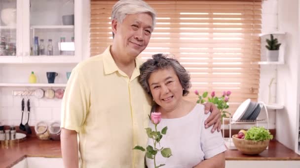 Asian elderly couple feeling happy smiling and holding flower and looking to camera while relax in kitchen at home. Lifestyle Senior family enjoy time at home concept. Portrait looking at camera. - Footage, Video