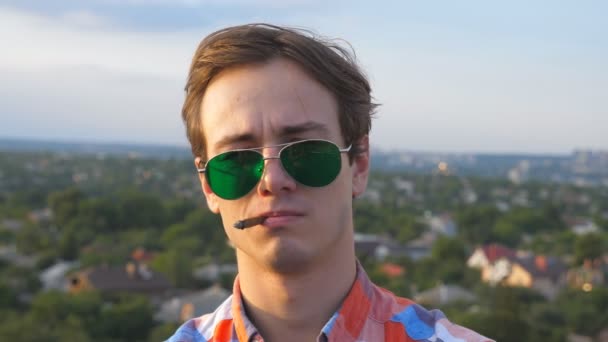 Handsome confident man in sunglasses standing on the edge of rooftop and smoking cigarette. Young serious guy resting on roof of high-rise building with cityscape at background. Dolly shot Close up - Séquence, vidéo