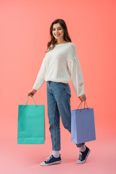 full length view of woman in white sweater and jeans holding shopping bags on pink background  - Foto, afbeelding