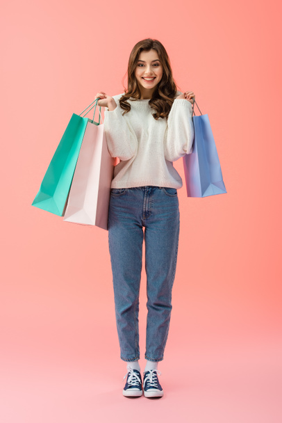 Full length view of smiling and attractive woman holding shopping bags on pink background  - Фото, зображення